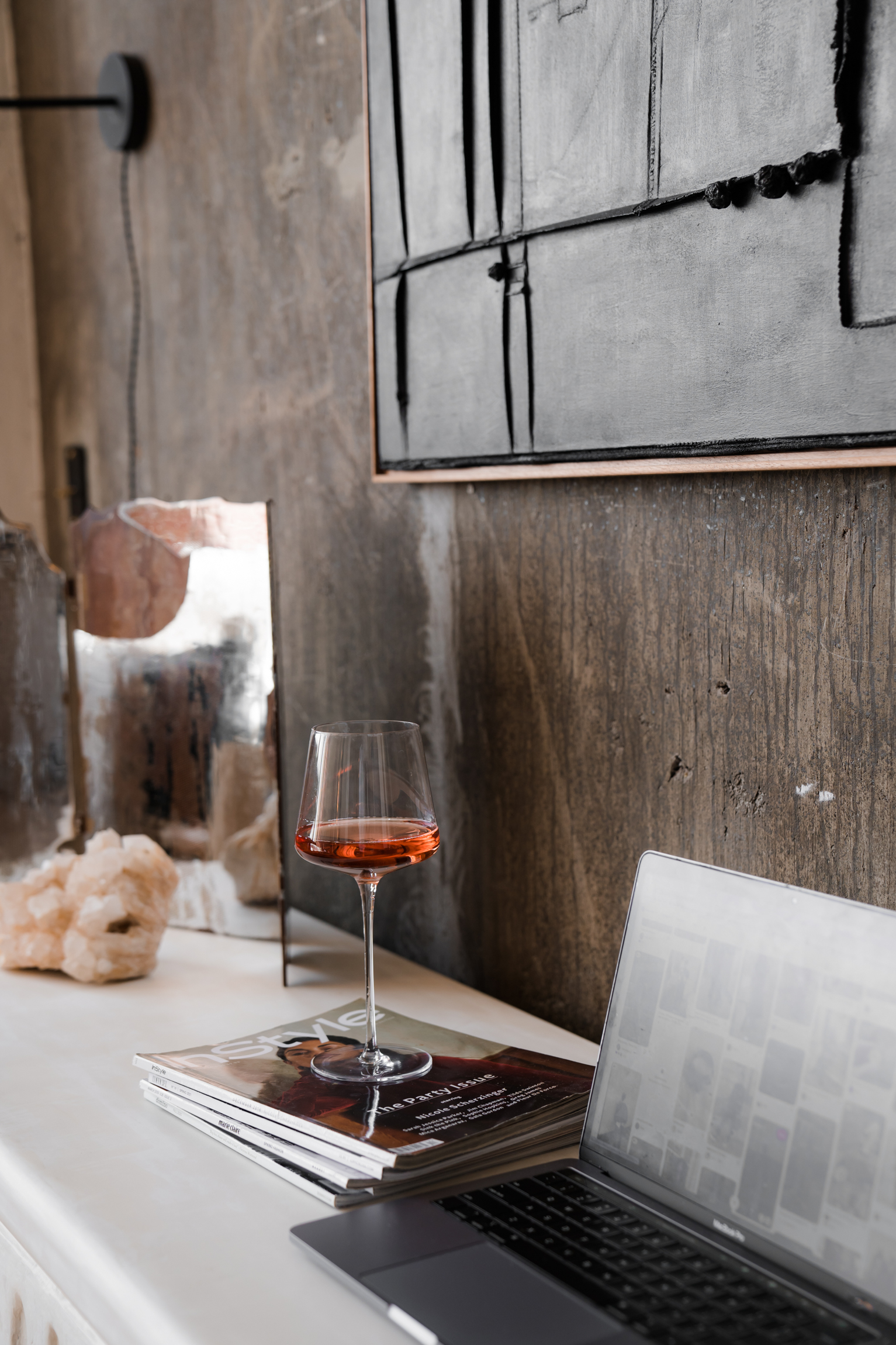 Moody workspace with a laptop, magazines, and a glass of rosé beside an abstract black and white painting.