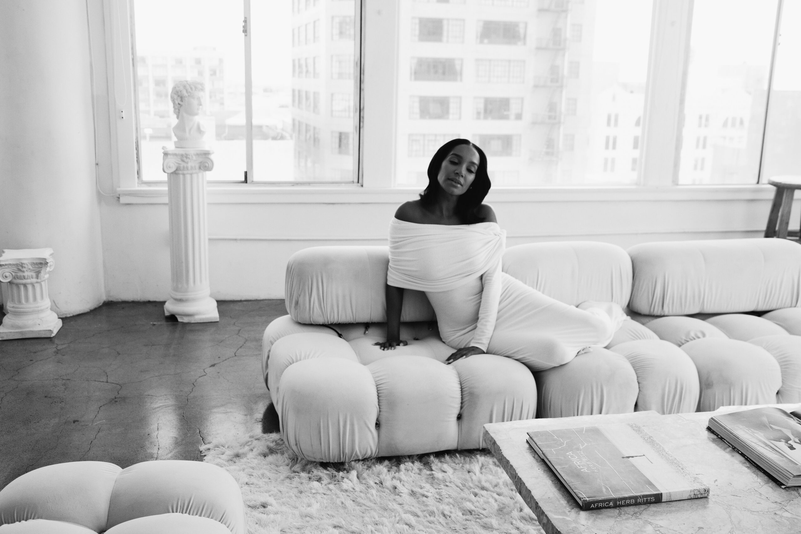 Black and white portrait of a woman seated on a white modular sofa in a bright, airy loft.